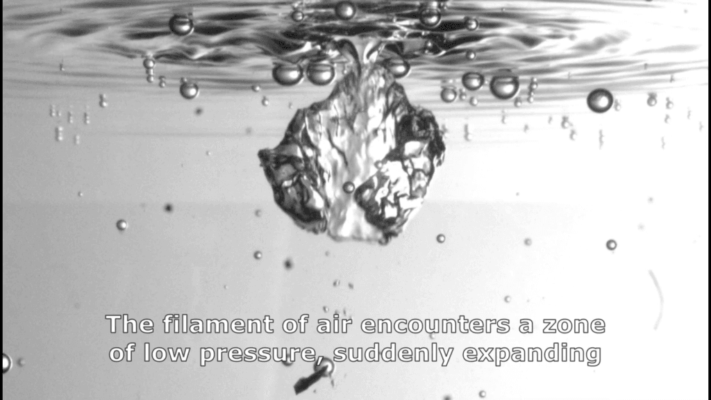 Black and white image showing a large, misshapen bubble in formation below the water surface. Text reads: &quot;The filament of air encounters a zone of low pressure, suddenly expanding&quot;