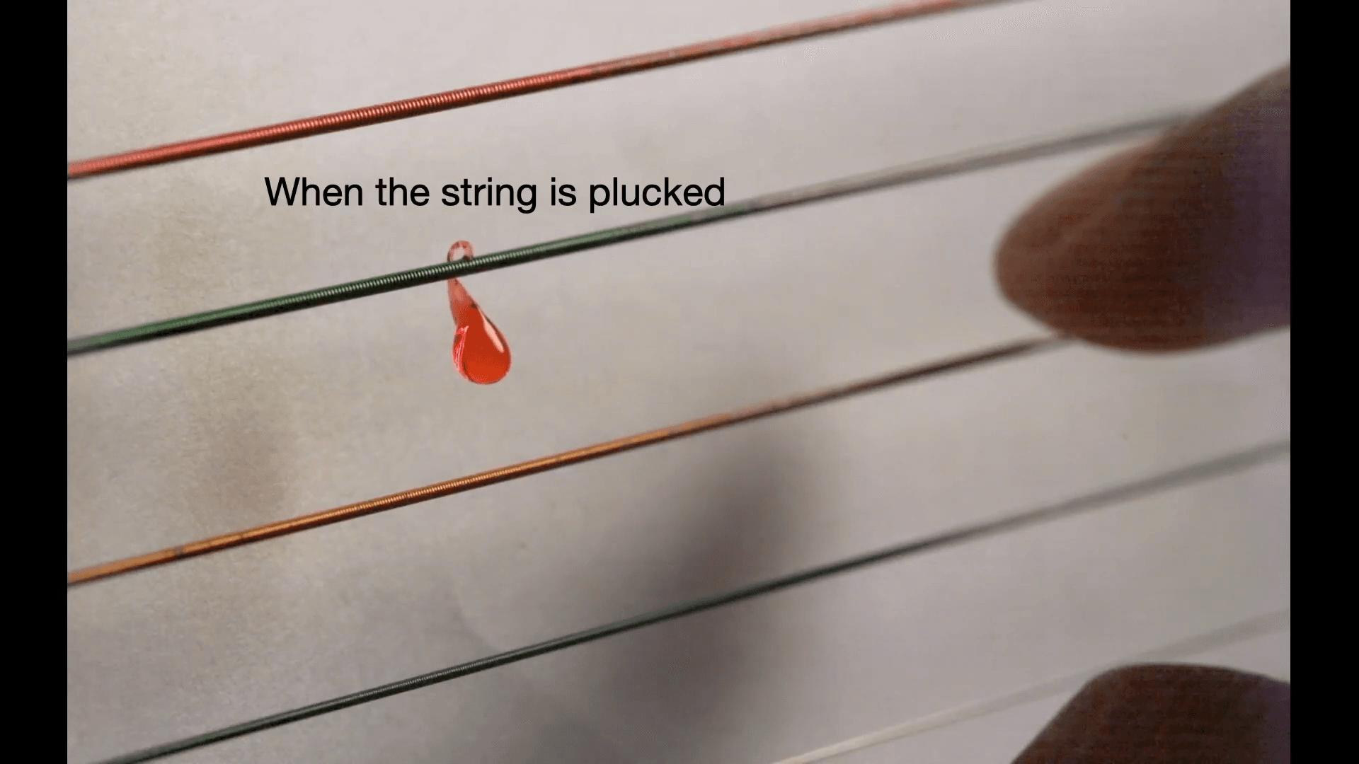 Close-up image of a red-dyed water droplet on a guitar string that's just been plucked. A blurry finger is just visible on the right side. Text reads: "When the string is plucked"