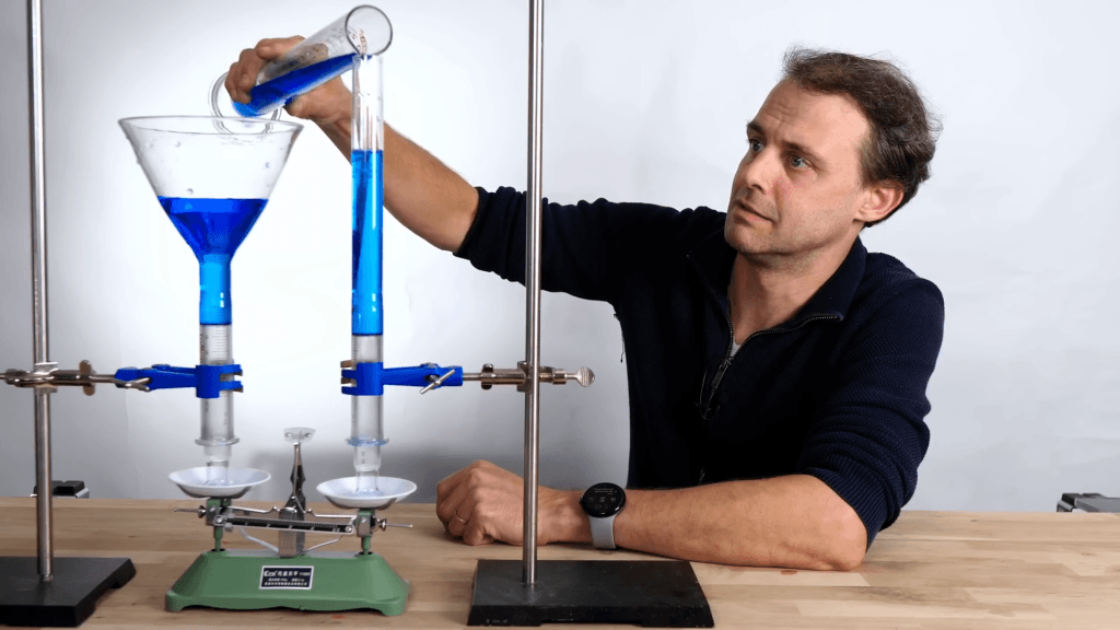 Image of Steve Mould pouring blue dyed water into a clear tube that sits on a scale, weighed against a second container with a different shape.