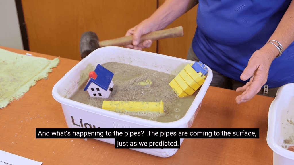 View into a white tub filled with wet sand, toy buildings, and toy pipes. A person holding a rubber mallet taps the tub and water pools at the surface of the sand, causing toys to rise to the surface and tip. Text reads, "And what's happening to the pipes? The pipes are coming to the surface, just as we predicted." View into a white tub filled with wet sand, toy buildings, and toy pipes. A person holding a rubber mallet taps the tub and water pools at the surface of the sand, causing toys to rise to the surface and tip. Text reads, "And what's happening to the pipes? The pipes are coming to the surface, just as we predicted."