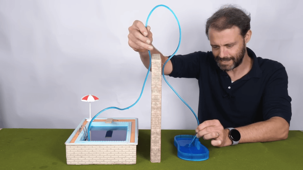 Steve Mould holds a miniature siphon running over a tabletop version of two swimming pools with a wall between them.